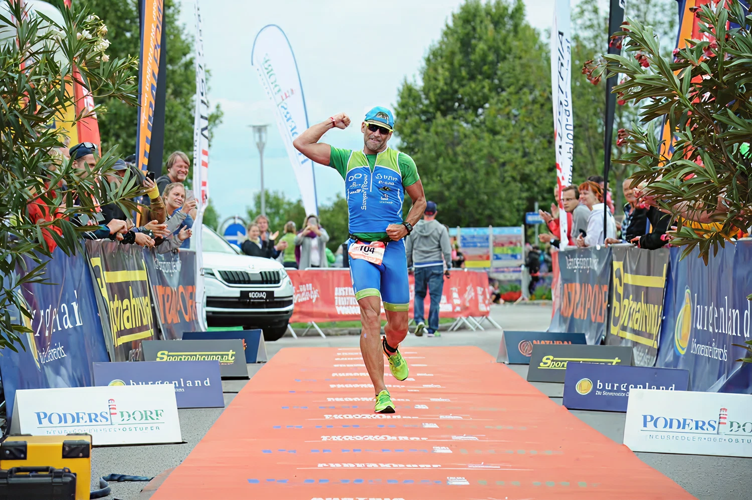 „Ein Triathlet jubelt beim Zieleinlauf, läuft über eine orange Ziellinie und hebt den Arm in die Höhe“